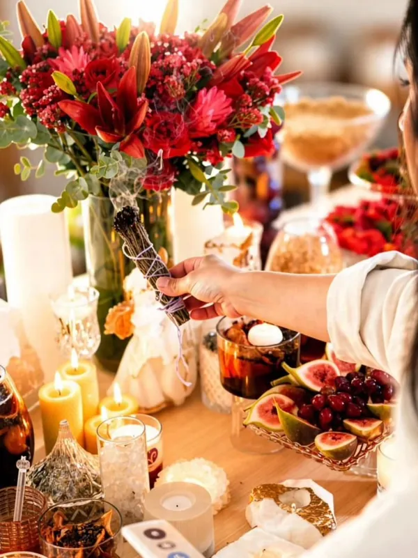 Mesa com flores vermelhas, frutas, velas e ervas, com um maço de ervas aceso
